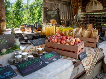 Rustic farm stand displaying wooden crates of fresh apples, glass jugs of juice, and paper bags. Chalkboard signs show products and prices.