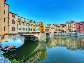 Ponte Vecchio bridge with colorful buildings spanning the Arno River in Florence under clear blue sky, with reflections in calm water.