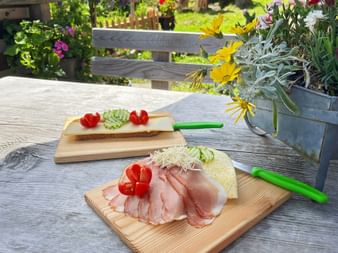 Wooden boards with ham, cheese, tomatoes and cucumber on a rustic table at Pitschenbergalm hut on Postalm, with flowers and garden in background.