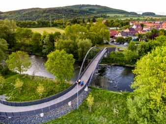 Luftaufnahme einer geschwungenen Fahrradbrücke mit Metallbogen über die Werra, umgeben von grünen Bäumen und einem Dorf mit Hügeln im Hintergrund.