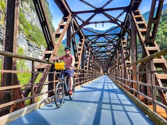 Radfahrer mit gelben Taschen auf historischer Metall-Fachwerkbrücke mit blauem Belag am Alpe-Adria-Radweg bei Chiusaforte, Berge sichtbar.
