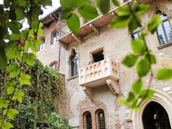 Historic stone balcony on medieval building in Verona, framed by green leaves. The famous Juliet's balcony features weathered brick walls.