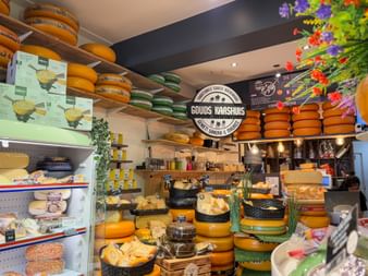 Innenansicht des Käseladens Gouds Kaashuis mit Regalen voller runder Gouda-Käselaibe in orangefarbenem Wachs, Kühlvitrine und Blumendekoration.