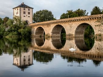 Historische Steinbrücke mit drei Bögen über die Lahn in Limburg. Ein weißer Schwan schwimmt im ruhigen Wasser, das Brücke und Turm spiegelt.