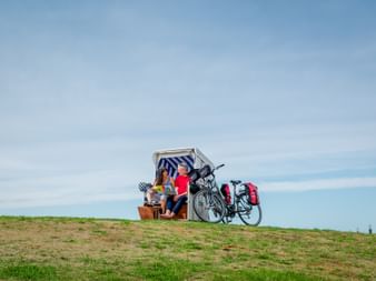 Zwei Radfahrer entspannen in einem weiß-blau gestreiften Strandkorb auf einem grasbewachsenen Deich. Zwei Fahrräder mit roten Taschen stehen daneben.