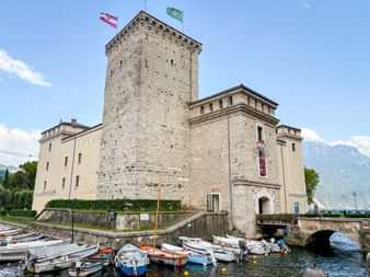 Medieval stone fortress Rocca di Riva with tower and flags, small boats moored in foreground, mountains visible in background under blue sky.
