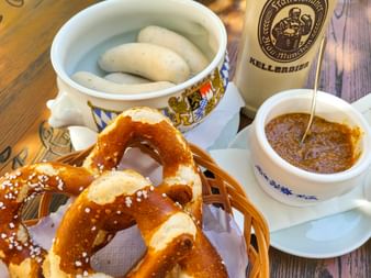 Traditional Bavarian meal with white sausages in ceramic bowl, pretzels with salt, mustard, and Kellerbier bottle on wooden table.
