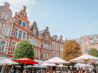 Historische flämische Giebelhäuser mit roten Backsteinfassaden säumen den Oude Markt in Löwen. Café-Terrassen mit Sonnenschirmen füllen den Platz.