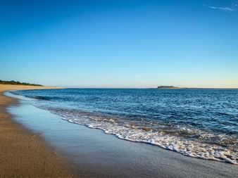 Sandstrand in Caminha mit sanften Wellen und blauem Wasser. Festung Forte da Ínsua auf kleiner Insel in der Ferne unter klarem Himmel.