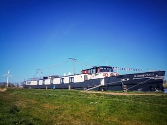 MS Magnifique IV, ein dunkelblau-weißes Flusskreuzfahrtschiff mit roten Akzenten, liegt an einem grasbewachsenen Wasserweg unter blauem Himmel.