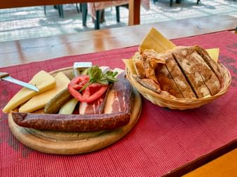 Wooden platter with sausage, cheese, ham, pickles, and tomatoes beside a basket of sliced bread on a red table mat.