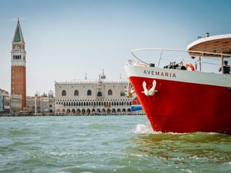 Red and white ship MS Ave Maria sailing in Venice lagoon with St. Mark's Campanile and Doge's Palace visible in the background under clear sky.