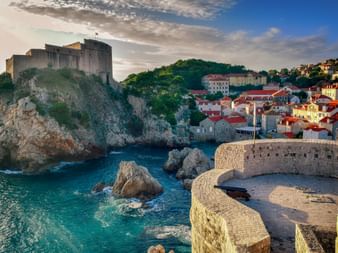 Fort Lovrijenac on rocky cliff overlooking turquoise Adriatic Sea in Dubrovnik, Croatia. Historic fortifications and red-roofed buildings visible.