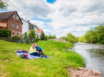 Two cyclists sitting on a blanket by a river near Lobenhausen. Bicycles parked on grass slope, half-timbered houses in background under cloudy sky.