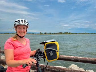 Lächelnde Radfahrerin mit weißem Helm und rosa Shirt steht mit Fahrrad an Holzgeländer an Lagune in Grado, Friaul.