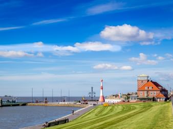 Red and white striped lighthouse at Bremerhaven harbor with historic brick buildings, green lawn, and boats in the water under blue sky.