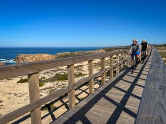 Zwei Radfahrer auf einem Holzsteg entlang der Küste in Nordportugal. Der Weg hat Geländer und verläuft neben Sandstrand und blauem Ozean.