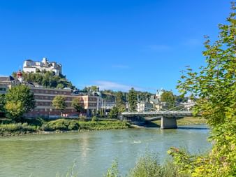 Festung Salzburg thront auf einem Hügel über der Salzach mit historischen Gebäuden, einer Brücke und üppiger grüner Vegetation unter blauem Himmel.