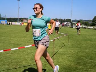 Female runner Nici in turquoise shirt and gray shorts crossing finish line at Grabenseelauf 2025 on grass field with spectators.