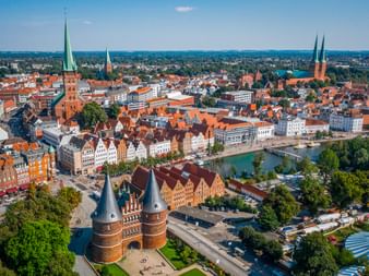 Luftaufnahme der Lübecker Altstadt mit roten Ziegeldächern, dem Holstentor im Vordergrund, Kirchtürmen und Uferpromenade entlang der Trave.