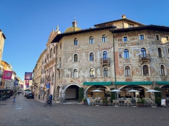 Historic frescoed buildings on Piazza del Duomo in Trient with outdoor café seating. A cyclist passes through the cobblestone square under clear blue sky.