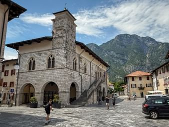 Historisches Rathaus aus Stein in Venzone mit Glockenturm und Arkadengang am Stadtplatz. Berge erheben sich im Hintergrund unter blauem Himmel.