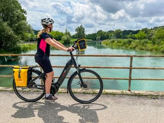 Radfahrerin mit Helm auf E-Bike mit gelben Taschen, pausiert auf Brücke über türkisfarbenem Kanal mit grüner Vegetation und bewölktem Himmel.