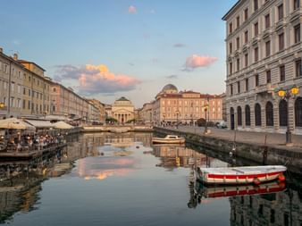 Canal Grande in Trieste with historic buildings, domed church, and boats moored in calm water reflecting the pink sunset sky.