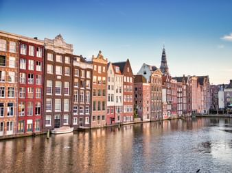 Reihe bunter traditioneller Giebelhäuser entlang des Damrak-Kanals in Amsterdam mit Kirchturm im Hintergrund unter blauem Himmel.