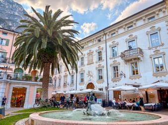 Fountain with water jets on a square in Riva del Garda, surrounded by historic buildings with ornate facades, palm tree, and outdoor dining.