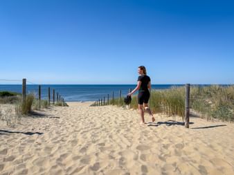 Frau in Radkleidung geht barfuß auf sandigem Weg zwischen Dünen zum Strand. Holzpfosten säumen den Weg mit Strandgras auf beiden Seiten.
