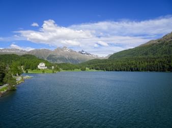 St. Moritz Lake surrounded by forested hills and snow-capped Alpine peaks. A white building sits on the green shoreline under blue sky.