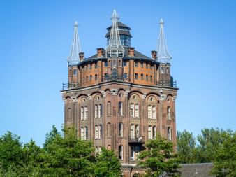 Historischer Backsteinwasserturm Villa Augustus in Dordrecht mit verzierter Architektur, weißen Türmchen und Aussichtsplattform umgeben von Bäumen.