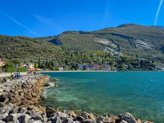 Rocky shoreline promenade in Torbole with turquoise Lake Garda waters, green mountains in the background, and colorful buildings along the coast.