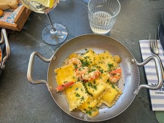 Metal pan with ravioli and scampi garnished with herbs, served with white wine and water on a gray table in Trieste.