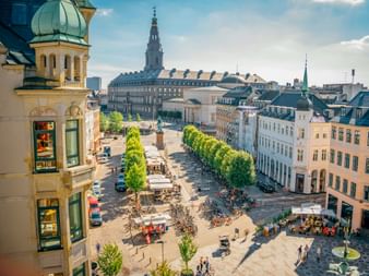 Luftaufnahme des Stadtplatzes von Malmö mit historischen Gebäuden, grünen Bäumen, Straßencafés und Radfahrern. Links ist eine Kirche mit grüner Kuppel.