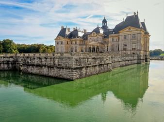 Baroque Château de Vaux-le-Vicomte surrounded by green water moat. The palace features cream stone facade with dark slate roofs and cupola.