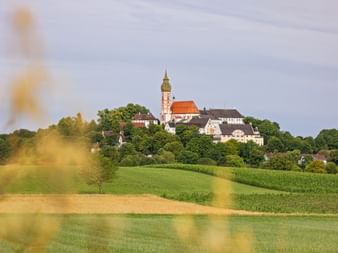 Andechs Monastery with distinctive tower and orange roof sits atop a green hill surrounded by trees and fields in Bavaria.