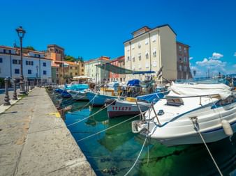 Boats moored in a marina along a stone promenade with colorful Mediterranean buildings under a clear blue sky.