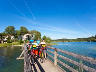 Vier Radfahrer mit Rädern auf einer Holzbrücke über einen See beim Kloster Seeon. Blauer Himmel und grüne Hügel im Hintergrund.
