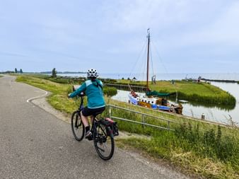 Radfahrer in türkisfarbener Jacke fährt entlang eines Deichwegs neben einem Kanal mit traditionellem Segelboot im grünen Marschland.