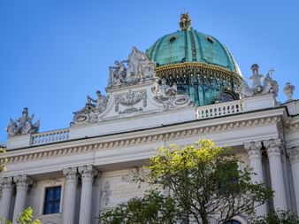 Green copper dome with golden ornaments atop the Hofburg Palace at Michaelerplatz in Vienna, featuring white sculptures and columns against blue sky.