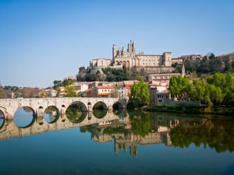 Stone bridge with arches crossing calm river in Béziers, with Cathedral Saint-Nazaire on hilltop above colorful houses under clear blue sky.