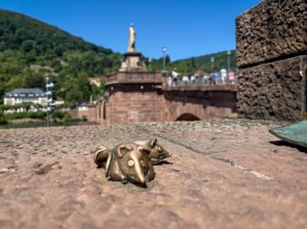 Kleine Bronzemaus-Skulptur auf Kopfsteinpflaster mit Heidelbergs Alter Brücke über dem Neckar im Hintergrund unter blauem Himmel.