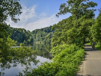 Two cyclists riding on paved path along the Ruhr river, surrounded by lush green trees and vegetation under partly cloudy sky.