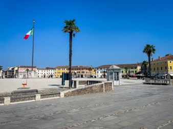 Wide view of Piazza Grande in Palmanova with Italian flag, palm trees, and colorful historic buildings under clear blue sky.