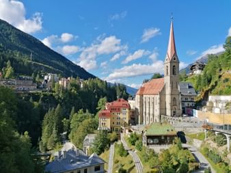 Church with tall copper spire in Bad Gastein surrounded by colorful buildings and green mountains under blue sky with white clouds.