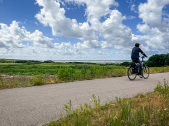 Radfahrer auf asphaltiertem Weg entlang des Weserradwegs mit grünen Wiesen und Wasserblick unter dramatischem Wolkenhimmel.