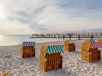 Colorful wicker beach chairs on sandy Baltic Sea beach with wooden pier extending into calm water under cloudy sky.