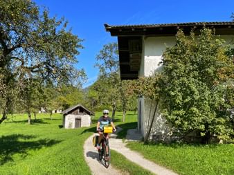 Female cyclist on gravel path between traditional Austrian farmhouse and green meadows with fruit trees under blue sky near Reit.